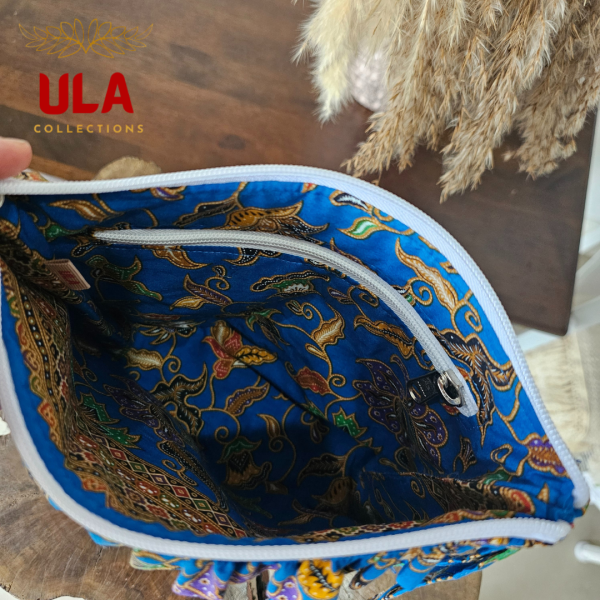 A vibrant zippered pouch bag or clutch with a solid royal blue cotton body. The bag is decorated with a wide, ruffled trim along the white zipper, made from a blue batik fabric with a multicolor floral print in shades of gold, purple, and red. The bag is shown open and includes a matching batik fabric wrist strap. It rests on a dark, rustic wooden surface, with a logo and dried pampas grass in the background, showcasing an eclectic fashion accessory perfect for travel or casual use.