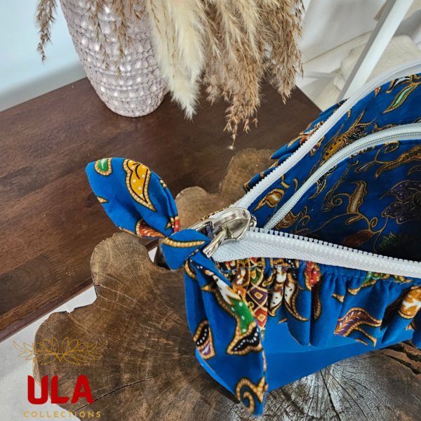A vibrant zippered pouch bag or clutch with a solid royal blue cotton body. The bag is decorated with a wide, ruffled trim along the white zipper, made from a blue batik fabric with a multicolor floral print in shades of gold, purple, and red. The bag is shown open and includes a matching batik fabric wrist strap. It rests on a dark, rustic wooden surface, with a logo and dried pampas grass in the background, showcasing an eclectic fashion accessory perfect for travel or casual use.