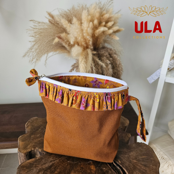 A stylish zippered pouch or clutch bag featuring a deep ochre brown cotton body with a subtle texture. The top is decorated with a wide, ruffled trim and interior lining made from a vibrant gold batik fabric with purple and brown floral motifs. This versatile fashion accessory includes a matching batik wrist strap and is shown styled as a handbag and resting on a wooden surface, offering an eclectic and bohemian accent for everyday or travel use.