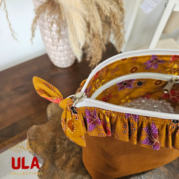 A stylish zippered pouch or clutch bag featuring a deep ochre brown cotton body with a subtle texture. The top is decorated with a wide, ruffled trim and interior lining made from a vibrant gold batik fabric with purple and brown floral motifs. This versatile fashion accessory includes a matching batik wrist strap and is shown styled as a handbag and resting on a wooden surface, offering an eclectic and bohemian accent for everyday or travel use.