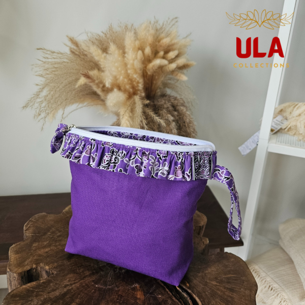 a square zippered pouch or clutch bag with a vibrant solid purple cotton body. The bag is accented with a ruffled trim along the white zipper, made from a black and white batik fabric featuring a floral or vine print. The bag also has a small wrist strap made from the same batik print fabric. This fashion accessory provides a bright pop of color and a bohemian or eclectic style, shown in a retail or closet setting.