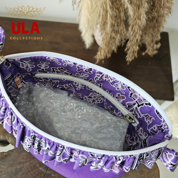 a square zippered pouch or clutch bag with a vibrant solid purple cotton body. The bag is accented with a ruffled trim along the white zipper, made from a black and white batik fabric featuring a floral or vine print. The bag also has a small wrist strap made from the same batik print fabric. This fashion accessory provides a bright pop of color and a bohemian or eclectic style, shown in a retail or closet setting.