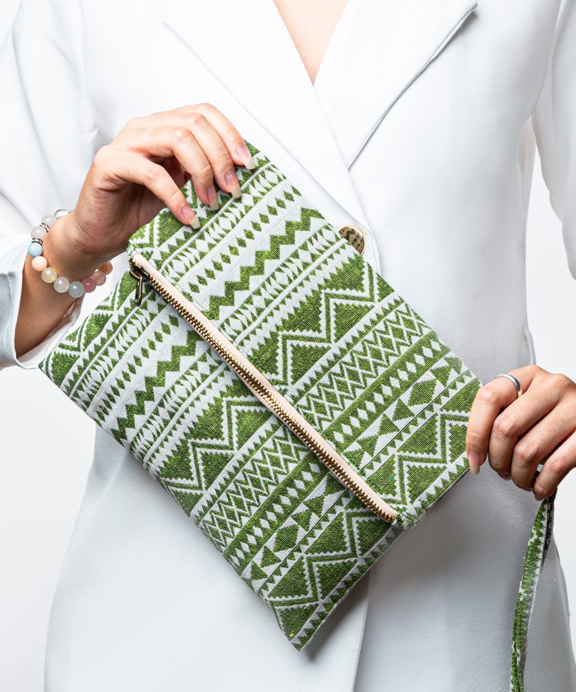A stylish fold-over clutch bag or zippered wristlet made from a textured woven fabric featuring an ethnic or tribal geometric pattern. The textile is colored in various shades of light and bright green, forming repeating vertical stripes of chevron and diamond motifs. The clutch has a decorative trim along the zipper line and a matching patterned fabric wrist strap. The green accessory is displayed against a white background with dried grasses, promoting a bohemian or eclectic fashion style.