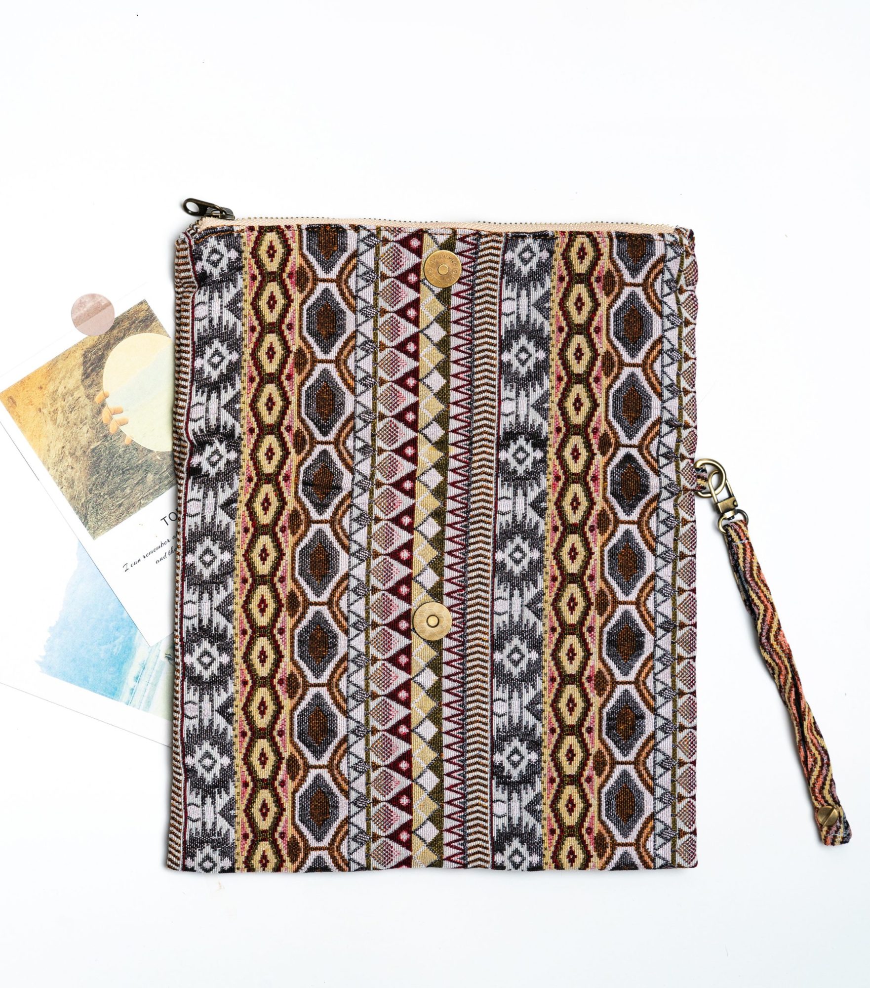 A stylish fold-over clutch bag or zippered wristlet made from a textured woven fabric featuring an intricate Southwestern or Aztec geometric pattern. The textile uses a rich, earthy color palette including shades of terracotta, dusty rose, brown, cream, and black. The clutch has a beige zigzag trim along the zipper line and a matching patterned fabric wrist strap. This bohemian fashion accessory is displayed on a white surface, ideal for everyday wear or travel.