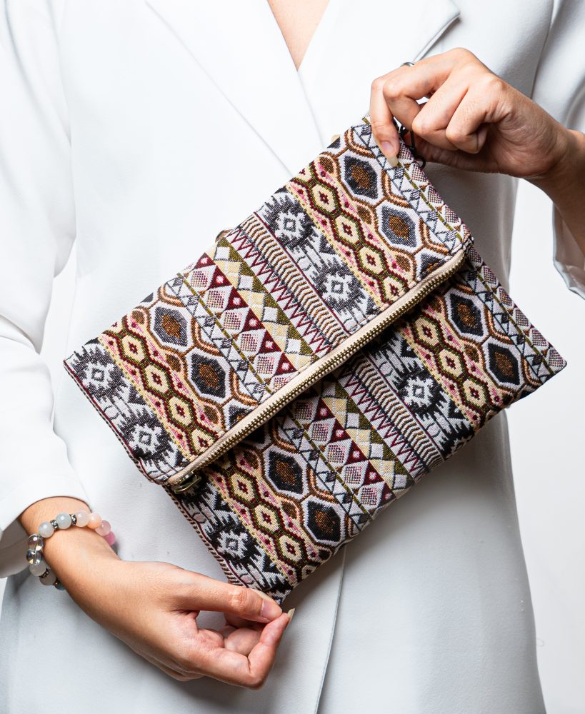 A stylish fold-over clutch bag or zippered wristlet made from a textured woven fabric featuring an intricate Southwestern or Aztec geometric pattern. The textile uses a rich, earthy color palette including shades of terracotta, dusty rose, brown, cream, and black. The clutch has a beige zigzag trim along the zipper line and a matching patterned fabric wrist strap. This bohemian fashion accessory is displayed on a white surface, ideal for everyday wear or travel.