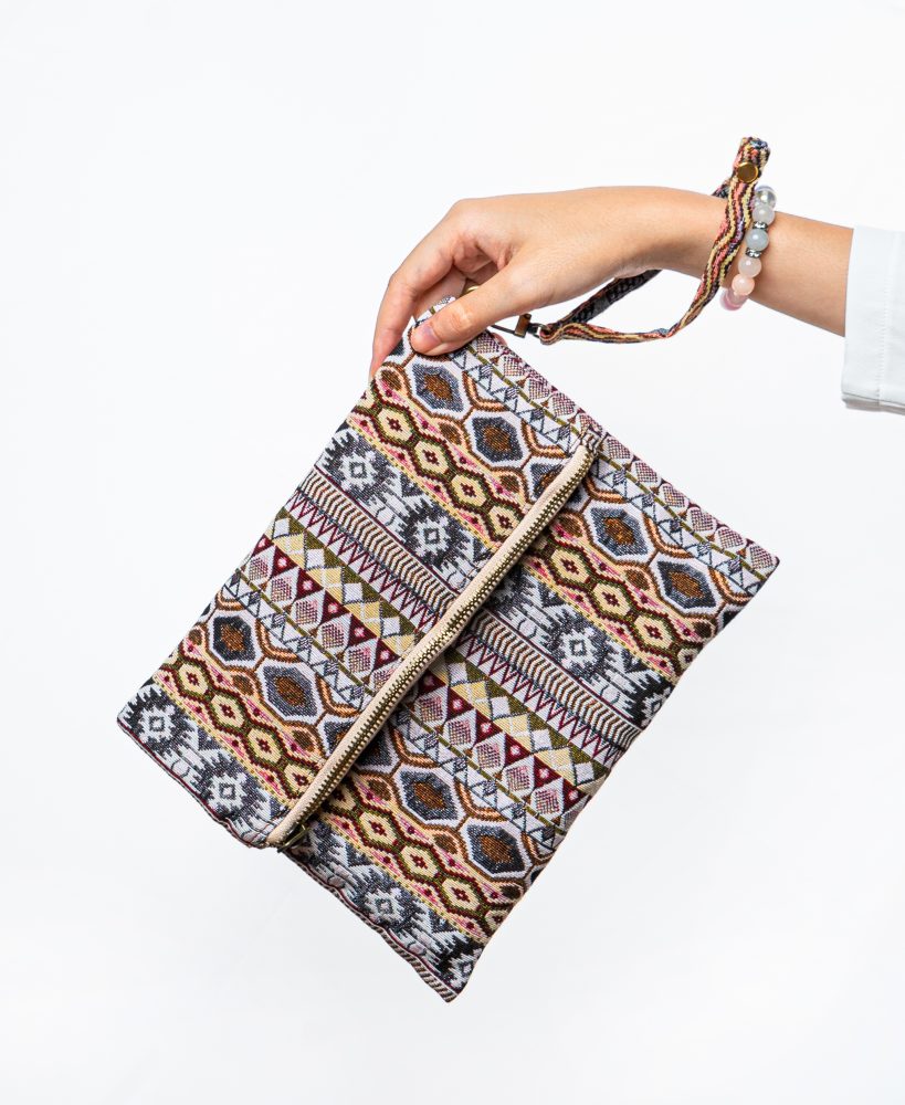 A stylish fold-over clutch bag or zippered wristlet made from a textured woven fabric featuring an intricate Southwestern or Aztec geometric pattern. The textile uses a rich, earthy color palette including shades of terracotta, dusty rose, brown, cream, and black. The clutch has a beige zigzag trim along the zipper line and a matching patterned fabric wrist strap. This bohemian fashion accessory is displayed on a white surface, ideal for everyday wear or travel.