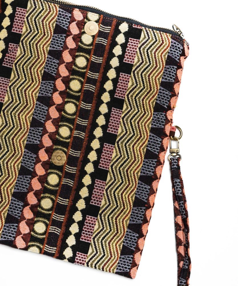 A stylish fold-over clutch bag or zippered wristlet made from a dark, textured woven fabric. The bag features an intricate African or Bohemian geometric pattern with vertical stripes in shades of black, dark brown, mustard yellow, burnt orange, and red. The clutch has a black zipper and a dark patterned fabric wrist strap. This maximalist fashion accessory is displayed on a magazine page, ideal for everyday use or adding an ethnic touch to an outfit.