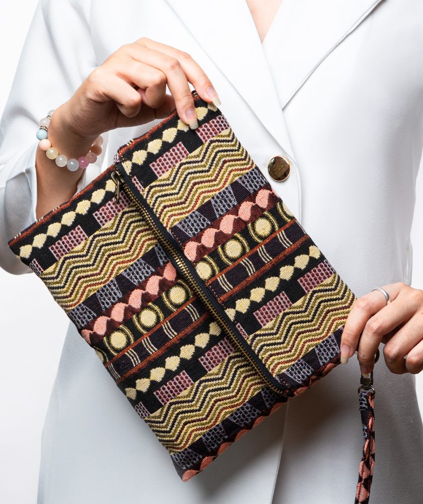A stylish fold-over clutch bag or zippered wristlet made from a dark, textured woven fabric. The bag features an intricate African or Bohemian geometric pattern with vertical stripes in shades of black, dark brown, mustard yellow, burnt orange, and red. The clutch has a black zipper and a dark patterned fabric wrist strap. This maximalist fashion accessory is displayed on a magazine page, ideal for everyday use or adding an ethnic touch to an outfit.