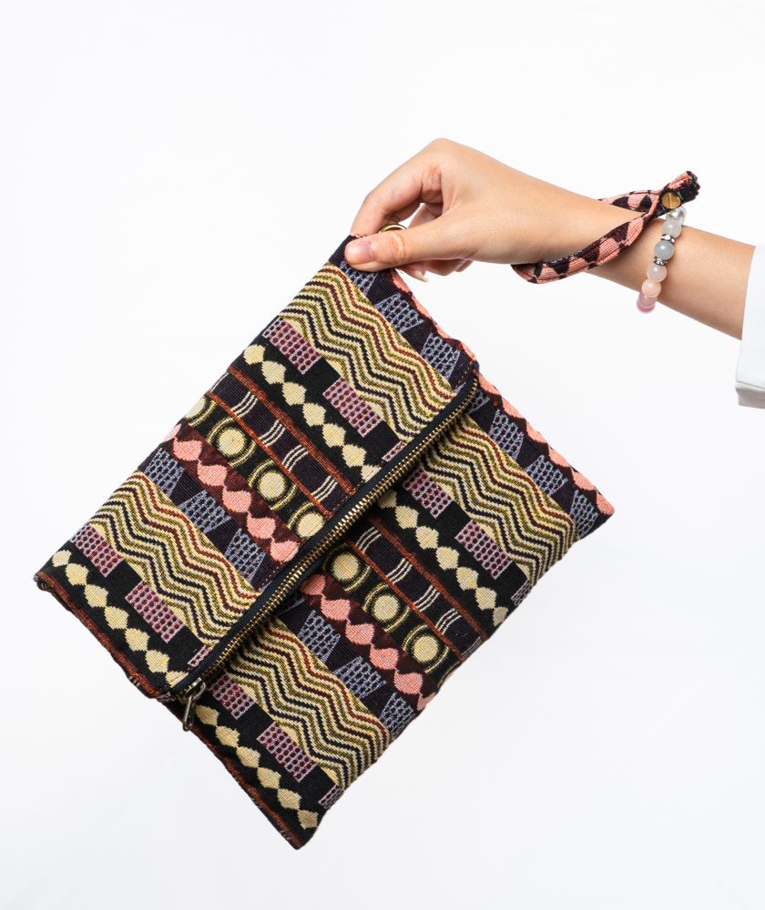 A stylish fold-over clutch bag or zippered wristlet made from a dark, textured woven fabric. The bag features an intricate African or Bohemian geometric pattern with vertical stripes in shades of black, dark brown, mustard yellow, burnt orange, and red. The clutch has a black zipper and a dark patterned fabric wrist strap. This maximalist fashion accessory is displayed on a magazine page, ideal for everyday use or adding an ethnic touch to an outfit.