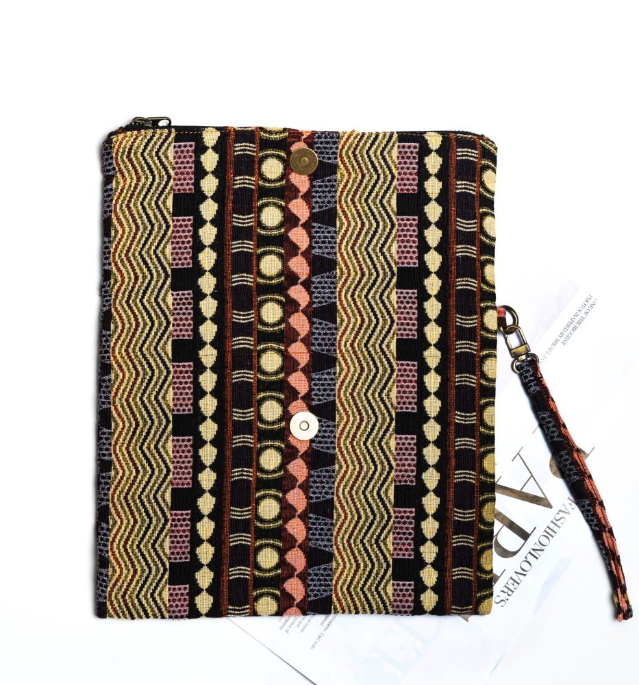 A stylish fold-over clutch bag or zippered wristlet made from a dark, textured woven fabric. The bag features an intricate African or Bohemian geometric pattern with vertical stripes in shades of black, dark brown, mustard yellow, burnt orange, and red. The clutch has a black zipper and a dark patterned fabric wrist strap. This maximalist fashion accessory is displayed on a magazine page, ideal for everyday use or adding an ethnic touch to an outfit.