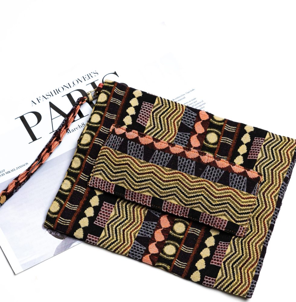 A stylish fold-over clutch bag or zippered wristlet made from a dark, textured woven fabric. The bag features an intricate African or Bohemian geometric pattern with vertical stripes in shades of black, dark brown, mustard yellow, burnt orange, and red. The clutch has a black zipper and a dark patterned fabric wrist strap. This maximalist fashion accessory is displayed on a magazine page, ideal for everyday use or adding an ethnic touch to an outfit.