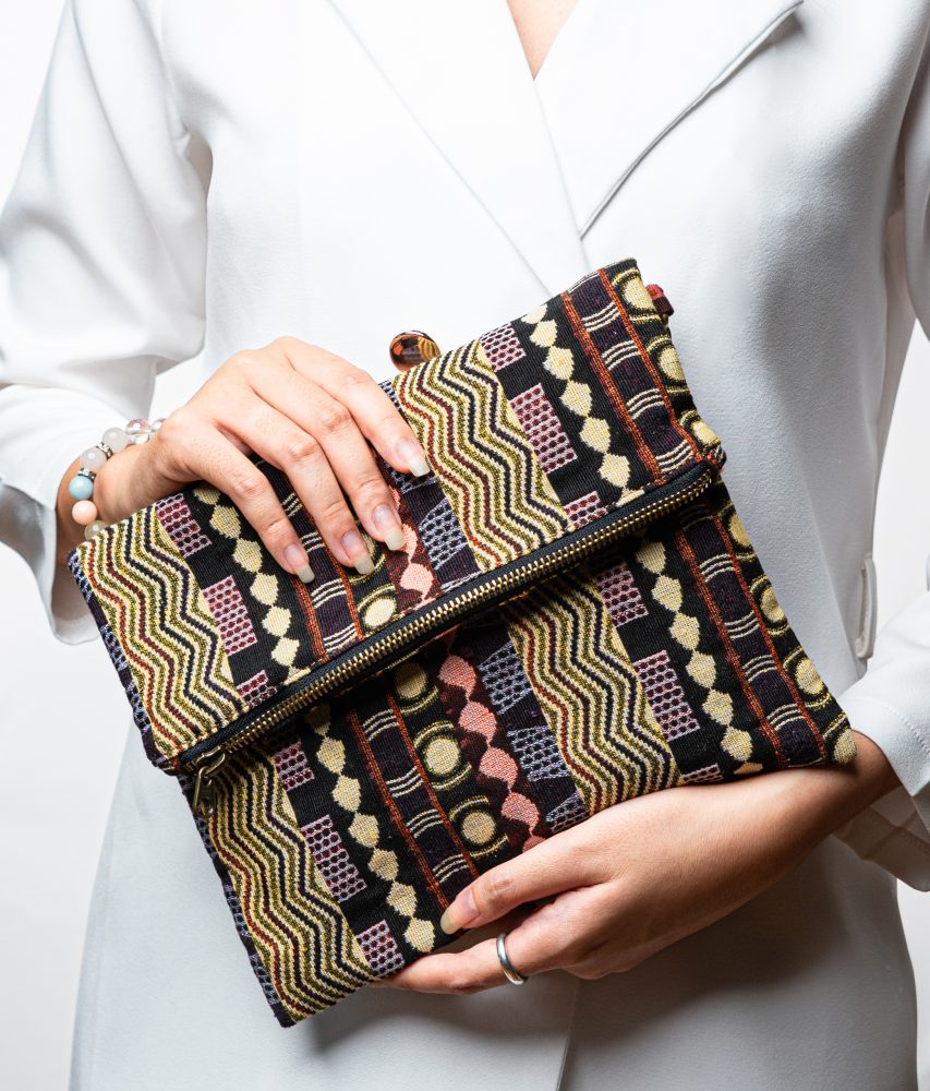 A stylish fold-over clutch bag or zippered wristlet made from a dark, textured woven fabric. The bag features an intricate African or Bohemian geometric pattern with vertical stripes in shades of black, dark brown, mustard yellow, burnt orange, and red. The clutch has a black zipper and a dark patterned fabric wrist strap. This maximalist fashion accessory is displayed on a magazine page, ideal for everyday use or adding an ethnic touch to an outfit.