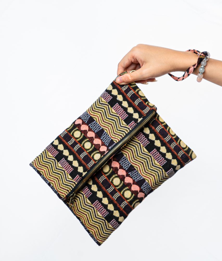 A stylish fold-over clutch bag or zippered wristlet made from a dark, textured woven fabric. The bag features an intricate African or Bohemian geometric pattern with vertical stripes in shades of black, dark brown, mustard yellow, burnt orange, and red. The clutch has a black zipper and a dark patterned fabric wrist strap. This maximalist fashion accessory is displayed on a magazine page, ideal for everyday use or adding an ethnic touch to an outfit.