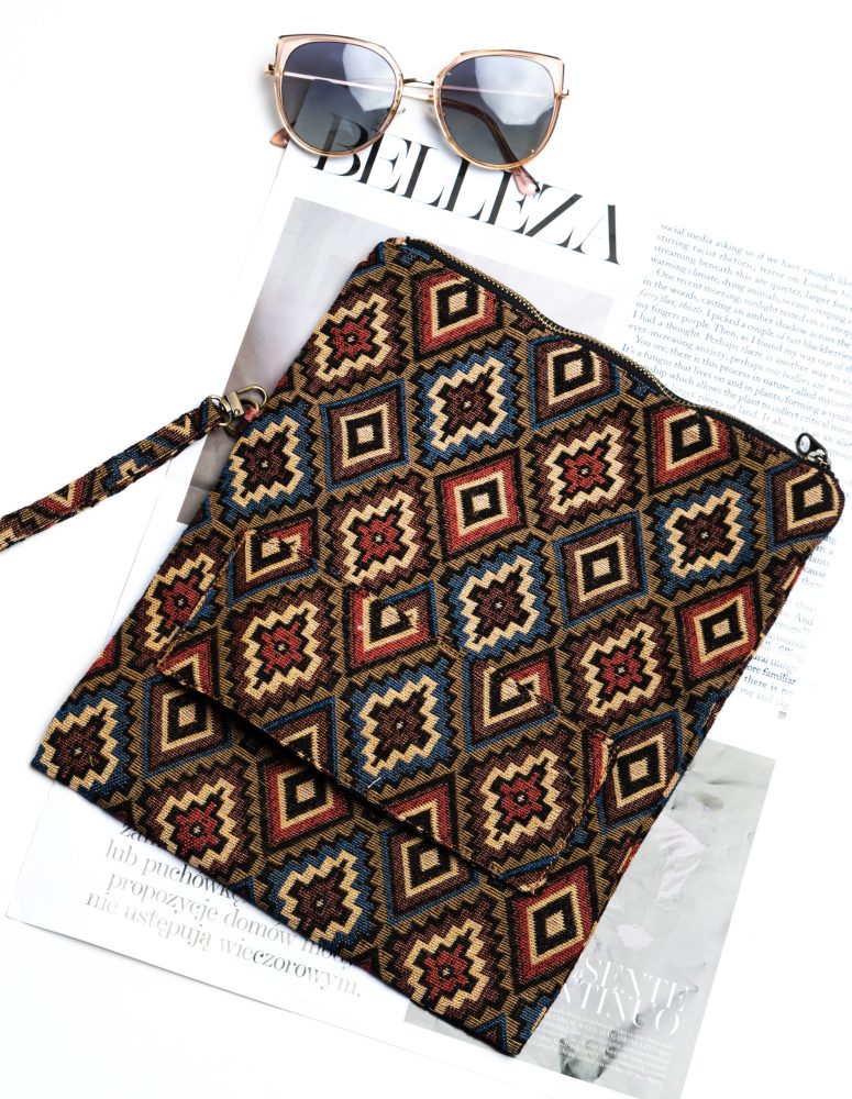 Here is the alt text for good SEO describing this image: A stylish fold-over clutch bag or zippered wristlet made from a dark, textured woven fabric. The bag features an intricate, repeating diamond geometric pattern in a rich, autumnal color palette including deep red, rust, light blue, and mustard yellow on a dark brown background. The clutch has a black zipper and a patterned fabric wrist strap. This bohemian fashion accessory is displayed on a magazine page, showcasing a maximalist or Southwestern style for everyday or travel use.