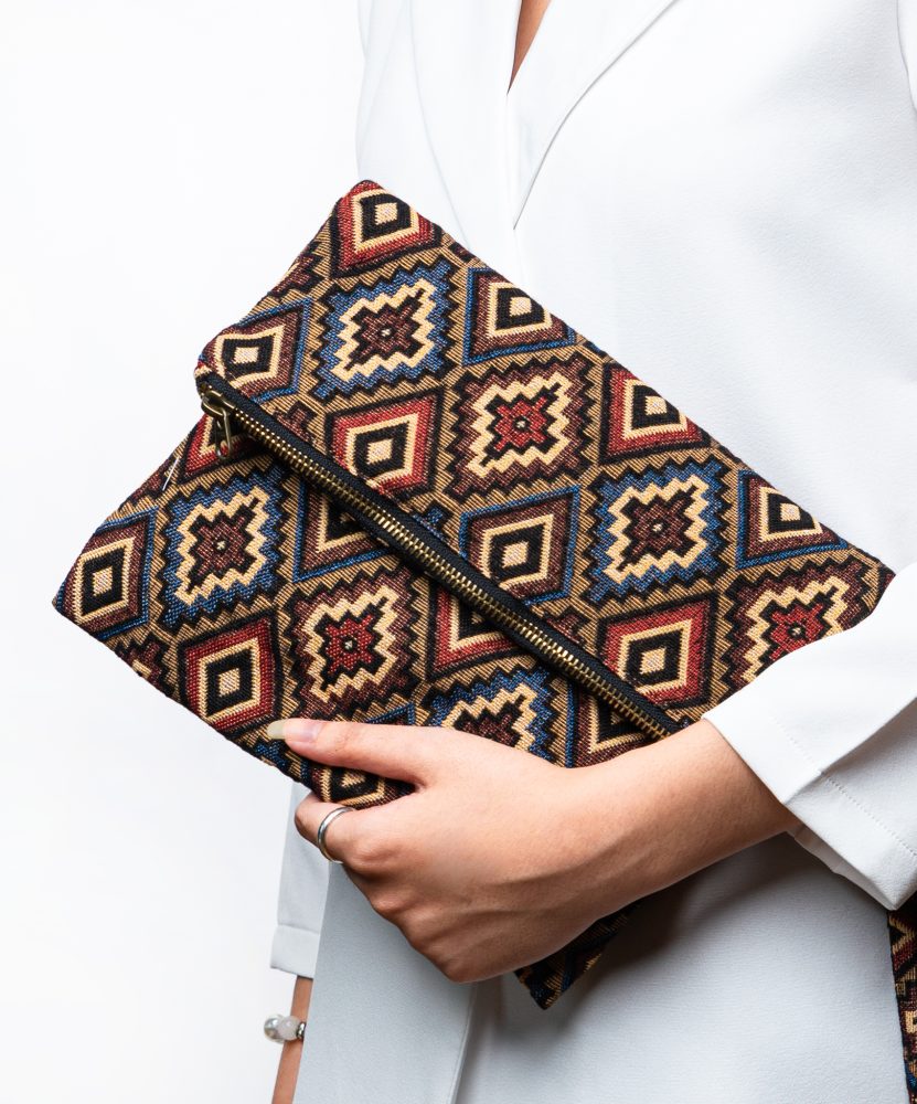 Here is the alt text for good SEO describing this image: A stylish fold-over clutch bag or zippered wristlet made from a dark, textured woven fabric. The bag features an intricate, repeating diamond geometric pattern in a rich, autumnal color palette including deep red, rust, light blue, and mustard yellow on a dark brown background. The clutch has a black zipper and a patterned fabric wrist strap. This bohemian fashion accessory is displayed on a magazine page, showcasing a maximalist or Southwestern style for everyday or travel use.