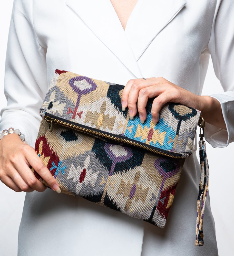 A stylish fold-over clutch bag or zippered wristlet made from a light-colored textured woven fabric. The bag features an intricate Southwestern or Ikat geometric pattern in muted, earthy tones including beige, gray, mustard yellow, dark teal, and burgundy. The clutch has a black zipper and a matching patterned fabric wrist strap. This bohemian fashion accessory is displayed on a magazine page, ideal for adding an eclectic tribal touch to an outfit for everyday wear or travel.