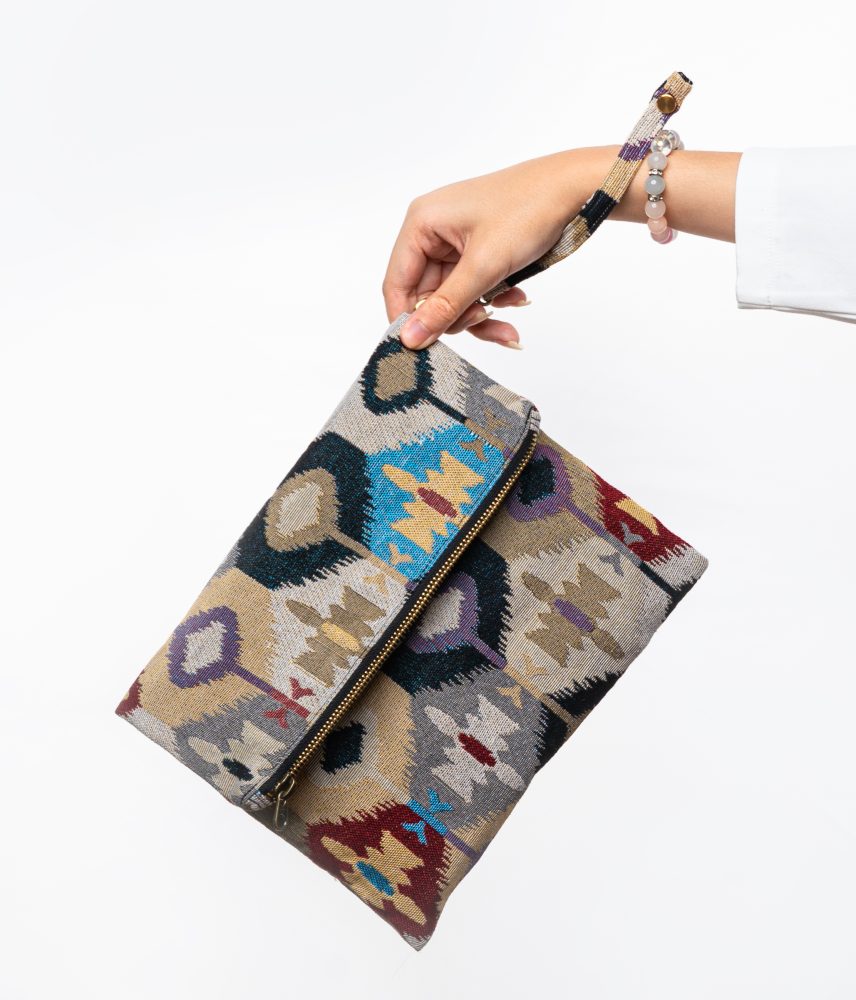 A stylish fold-over clutch bag or zippered wristlet made from a light-colored textured woven fabric. The bag features an intricate Southwestern or Ikat geometric pattern in muted, earthy tones including beige, gray, mustard yellow, dark teal, and burgundy. The clutch has a black zipper and a matching patterned fabric wrist strap. This bohemian fashion accessory is displayed on a magazine page, ideal for adding an eclectic tribal touch to an outfit for everyday wear or travel.