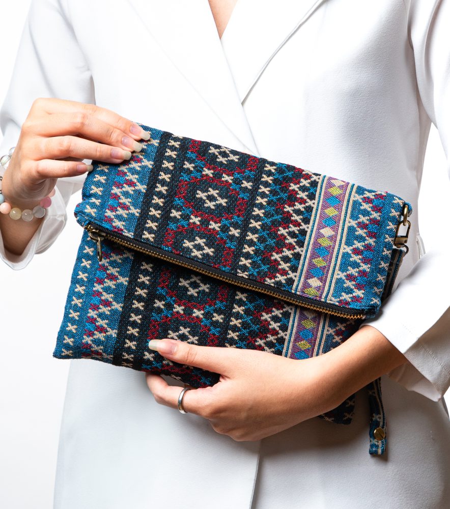 A stylish fold-over clutch bag or zippered wristlet made from a textured woven fabric featuring an ethnic or Aztec geometric pattern. The textile is primarily in shades of teal, deep blue, and cream, accented with dark burgundy, black, and gold woven details. The clutch has a metal zipper and a matching patterned fabric wrist strap, and is displayed against a white background, promoting it as a bohemian fashion accessory or travel handbag.