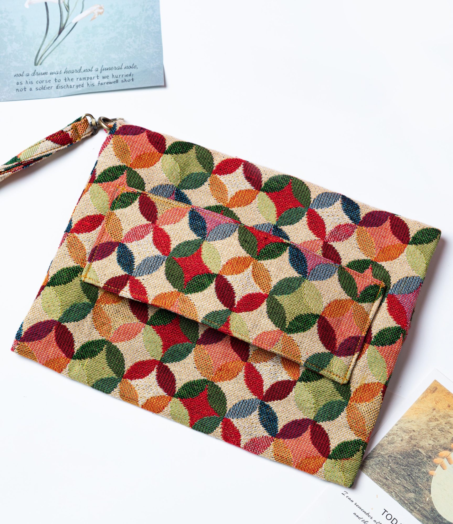 A stylish fold-over clutch bag or zippered wristlet made from a textured natural woven fabric. The bag features a vibrant, multi-color geometric pattern of interlocking circles or Seigaiha wave motifs in shades of red, teal, orange, green, and pink. The clutch has a white zipper and a matching patterned fabric wrist strap, and is displayed on a white surface with a cork coaster and wicker mat in the background, promoting it as a bohemian fashion accessory.