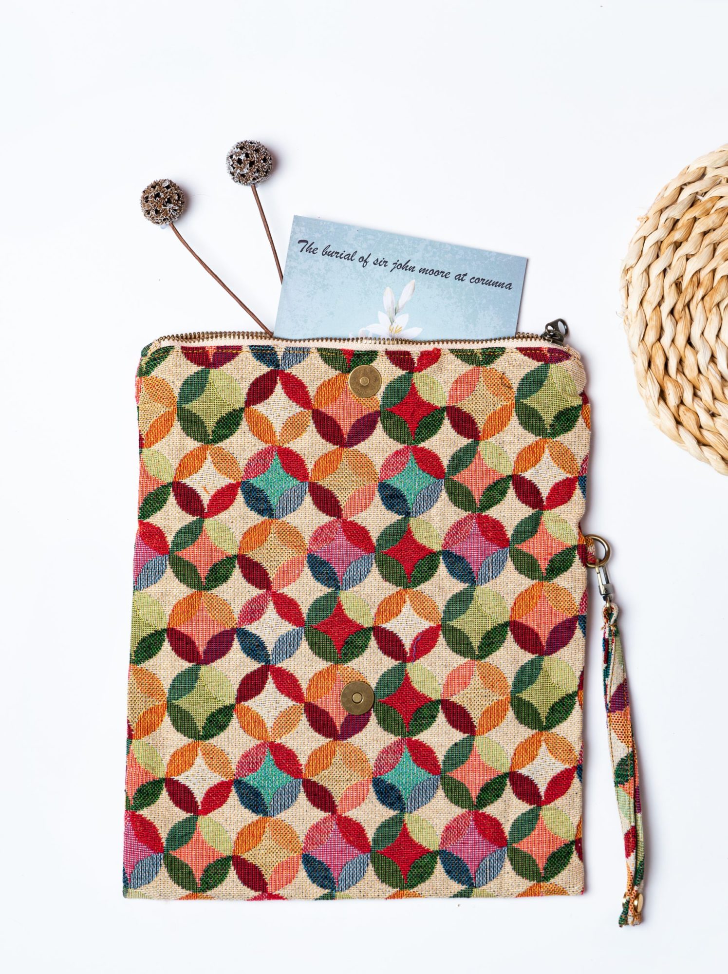 A stylish fold-over clutch bag or zippered wristlet made from a textured natural woven fabric. The bag features a vibrant, multi-color geometric pattern of interlocking circles or Seigaiha wave motifs in shades of red, teal, orange, green, and pink. The clutch has a white zipper and a matching patterned fabric wrist strap, and is displayed on a white surface with a cork coaster and wicker mat in the background, promoting it as a bohemian fashion accessory.