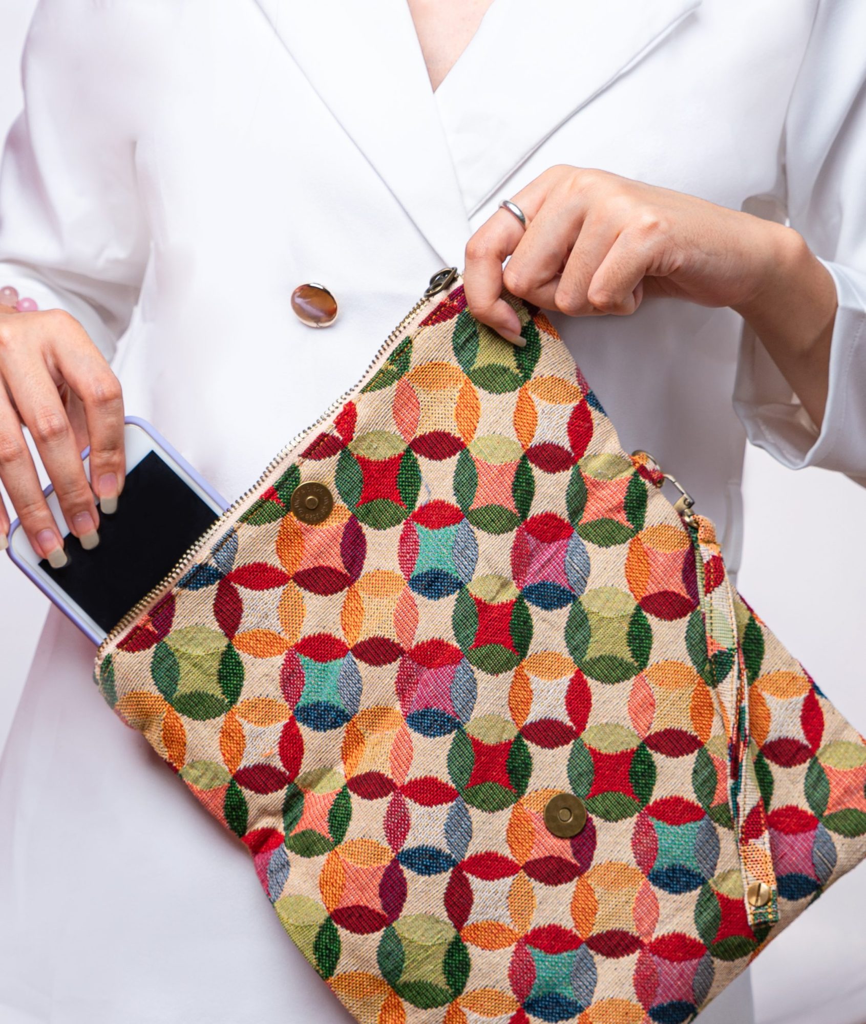 A stylish fold-over clutch bag or zippered wristlet made from a textured natural woven fabric. The bag features a vibrant, multi-color geometric pattern of interlocking circles or Seigaiha wave motifs in shades of red, teal, orange, green, and pink. The clutch has a white zipper and a matching patterned fabric wrist strap, and is displayed on a white surface with a cork coaster and wicker mat in the background, promoting it as a bohemian fashion accessory.