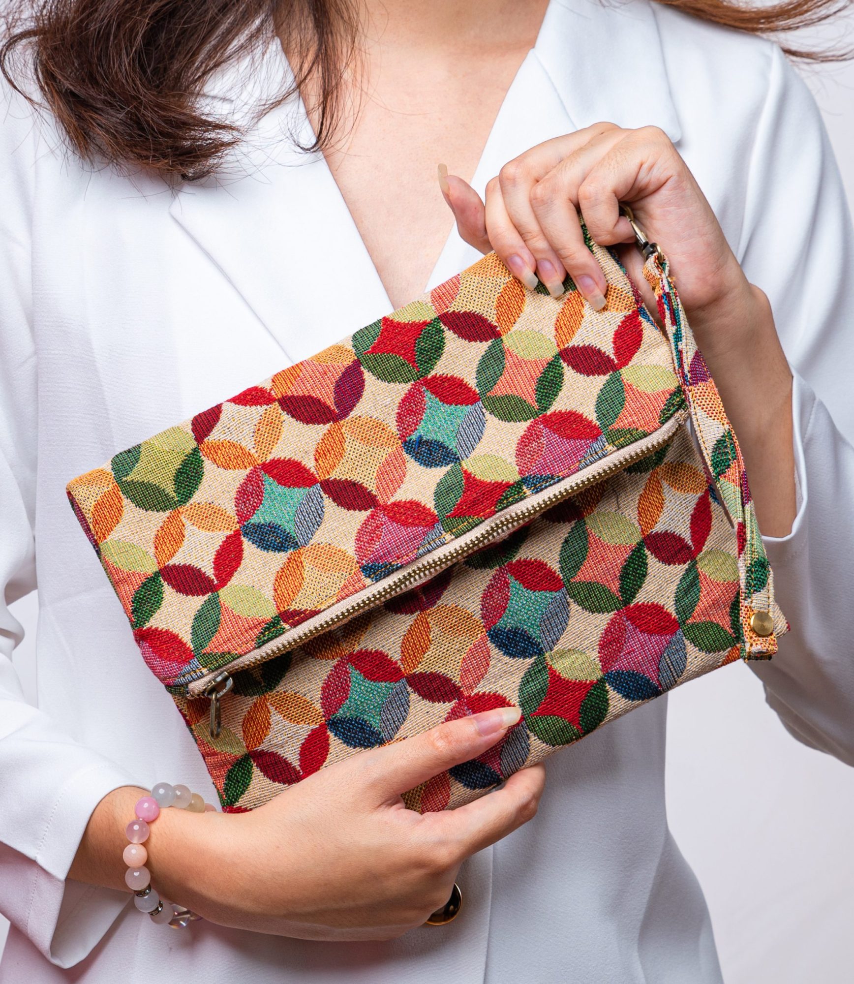 A stylish fold-over clutch bag or zippered wristlet made from a textured natural woven fabric. The bag features a vibrant, multi-color geometric pattern of interlocking circles or Seigaiha wave motifs in shades of red, teal, orange, green, and pink. The clutch has a white zipper and a matching patterned fabric wrist strap, and is displayed on a white surface with a cork coaster and wicker mat in the background, promoting it as a bohemian fashion accessory.