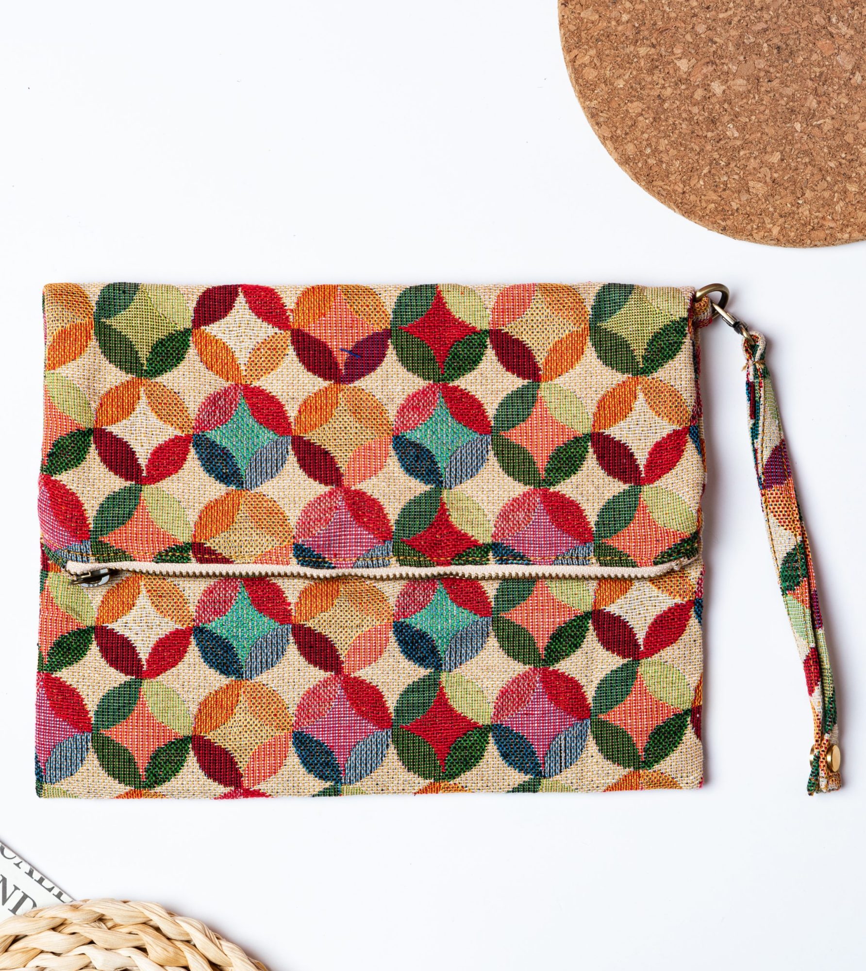 A stylish fold-over clutch bag or zippered wristlet made from a textured natural woven fabric. The bag features a vibrant, multi-color geometric pattern of interlocking circles or Seigaiha wave motifs in shades of red, teal, orange, green, and pink. The clutch has a white zipper and a matching patterned fabric wrist strap, and is displayed on a white surface with a cork coaster and wicker mat in the background, promoting it as a bohemian fashion accessory.