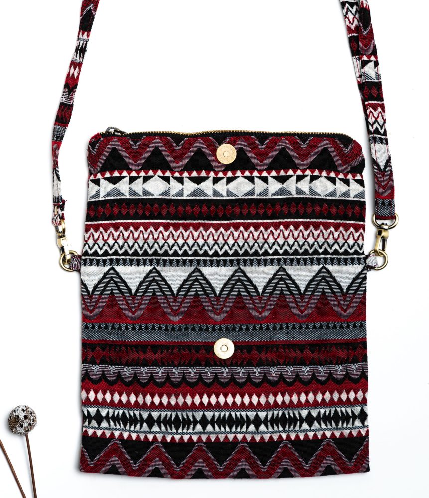 A stylish fold-over crossbody bag or clutch made from a textured woven fabric. The bag features an intricate tribal or geometric pattern with repeating chevron and diamond motifs in a high-contrast color palette of red, black, white, and gray. The accessory has a black zipper along the fold and a detachable, matching patterned fabric strap with gold-toned hardware, making it a versatile bohemian fashion accessory for everyday wear or travel.