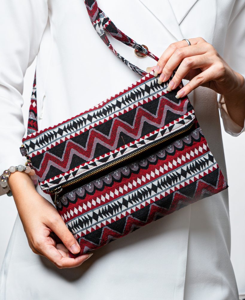 A stylish fold-over crossbody bag or clutch made from a textured woven fabric. The bag features an intricate tribal or geometric pattern with repeating chevron and diamond motifs in a high-contrast color palette of red, black, white, and gray. The accessory has a black zipper along the fold and a detachable, matching patterned fabric strap with gold-toned hardware, making it a versatile bohemian fashion accessory for everyday wear or travel.