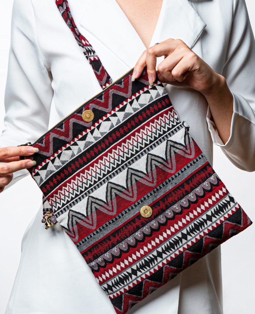 A stylish fold-over crossbody bag or clutch made from a textured woven fabric. The bag features an intricate tribal or geometric pattern with repeating chevron and diamond motifs in a high-contrast color palette of red, black, white, and gray. The accessory has a black zipper along the fold and a detachable, matching patterned fabric strap with gold-toned hardware, making it a versatile bohemian fashion accessory for everyday wear or travel.