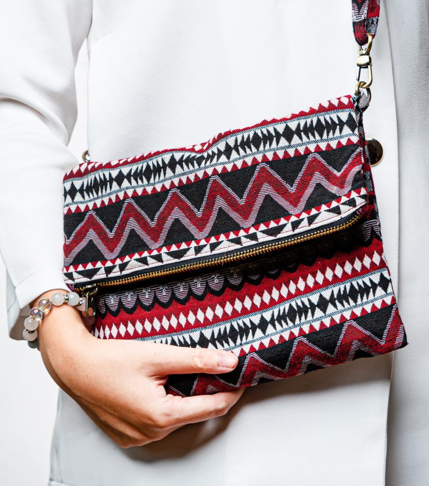 A stylish fold-over crossbody bag or clutch made from a textured woven fabric. The bag features an intricate tribal or geometric pattern with repeating chevron and diamond motifs in a high-contrast color palette of red, black, white, and gray. The accessory has a black zipper along the fold and a detachable, matching patterned fabric strap with gold-toned hardware, making it a versatile bohemian fashion accessory for everyday wear or travel.