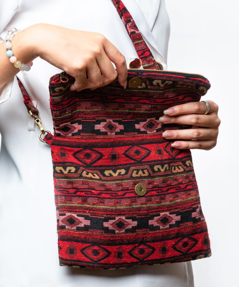 A person's hand is holding up a fold-over crossbody bag or shoulder bag made from a dark, textured woven fabric. The bag features an intricate tribal or geometric pattern with repeating motifs in a rich color palette of red, burgundy, black, and gold. The bag has a black zipper along the fold and a wide, detachable, matching patterned fabric strap with gold-toned hardware, showcasing a bohemian fashion accessory for everyday wear or travel.