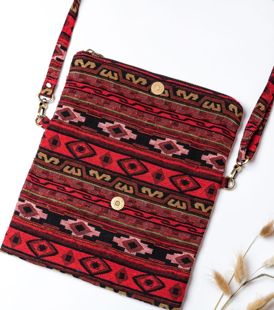 A person's hand is holding up a fold-over crossbody bag or shoulder bag made from a dark, textured woven fabric. The bag features an intricate tribal or geometric pattern with repeating motifs in a rich color palette of red, burgundy, black, and gold. The bag has a black zipper along the fold and a wide, detachable, matching patterned fabric strap with gold-toned hardware, showcasing a bohemian fashion accessory for everyday wear or travel.