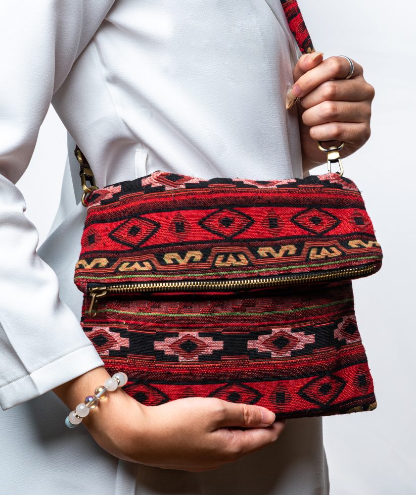 A person's hand is holding up a fold-over crossbody bag or shoulder bag made from a dark, textured woven fabric. The bag features an intricate tribal or geometric pattern with repeating motifs in a rich color palette of red, burgundy, black, and gold. The bag has a black zipper along the fold and a wide, detachable, matching patterned fabric strap with gold-toned hardware, showcasing a bohemian fashion accessory for everyday wear or travel.