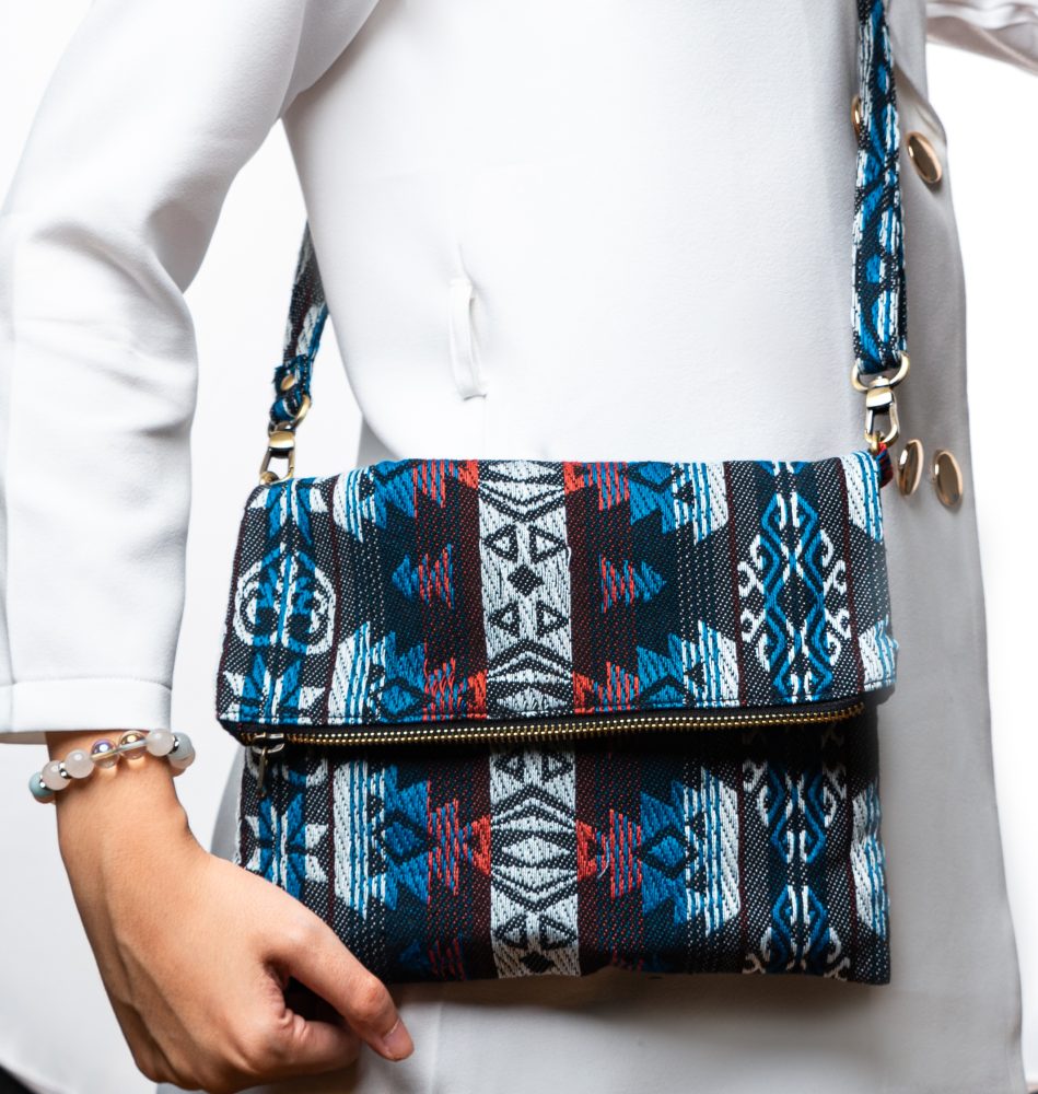 A stylish fold-over crossbody bag or shoulder bag made from a dark woven fabric. The bag features an intricate tribal or Ikat geometric pattern in a dramatic color palette of black, deep indigo, turquoise, dark red, and white. The bag has a zipper along the fold and a wide, matching patterned fabric strap with silver-toned hardware, showcasing a maximalist or eclectic fashion accessory for everyday wear or travel.