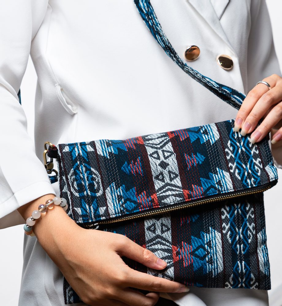 A stylish fold-over crossbody bag or shoulder bag made from a dark woven fabric. The bag features an intricate tribal or Ikat geometric pattern in a dramatic color palette of black, deep indigo, turquoise, dark red, and white. The bag has a zipper along the fold and a wide, matching patterned fabric strap with silver-toned hardware, showcasing a maximalist or eclectic fashion accessory for everyday wear or travel.