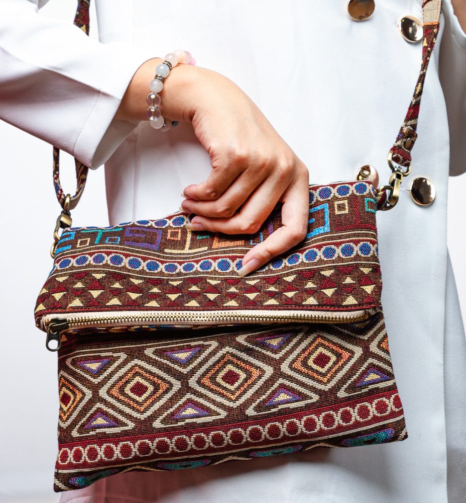 A stylish fold-over crossbody bag or shoulder bag made from a dark brown woven fabric. The bag features an intricate Southwestern or Aztec geometric pattern with horizontal stripes of diamond, zigzag, and Greek key motifs in shades of burgundy, deep blue, turquoise, and orange. The bag has a small zipper along the fold and a wide, matching patterned fabric strap with silver-toned hardware, showcasing an eclectic fashion accessory for everyday wear or travel.