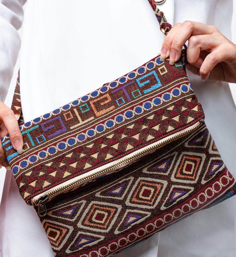 A stylish fold-over crossbody bag or shoulder bag made from a dark brown woven fabric. The bag features an intricate Southwestern or Aztec geometric pattern with horizontal stripes of diamond, zigzag, and Greek key motifs in shades of burgundy, deep blue, turquoise, and orange. The bag has a small zipper along the fold and a wide, matching patterned fabric strap with silver-toned hardware, showcasing an eclectic fashion accessory for everyday wear or travel.