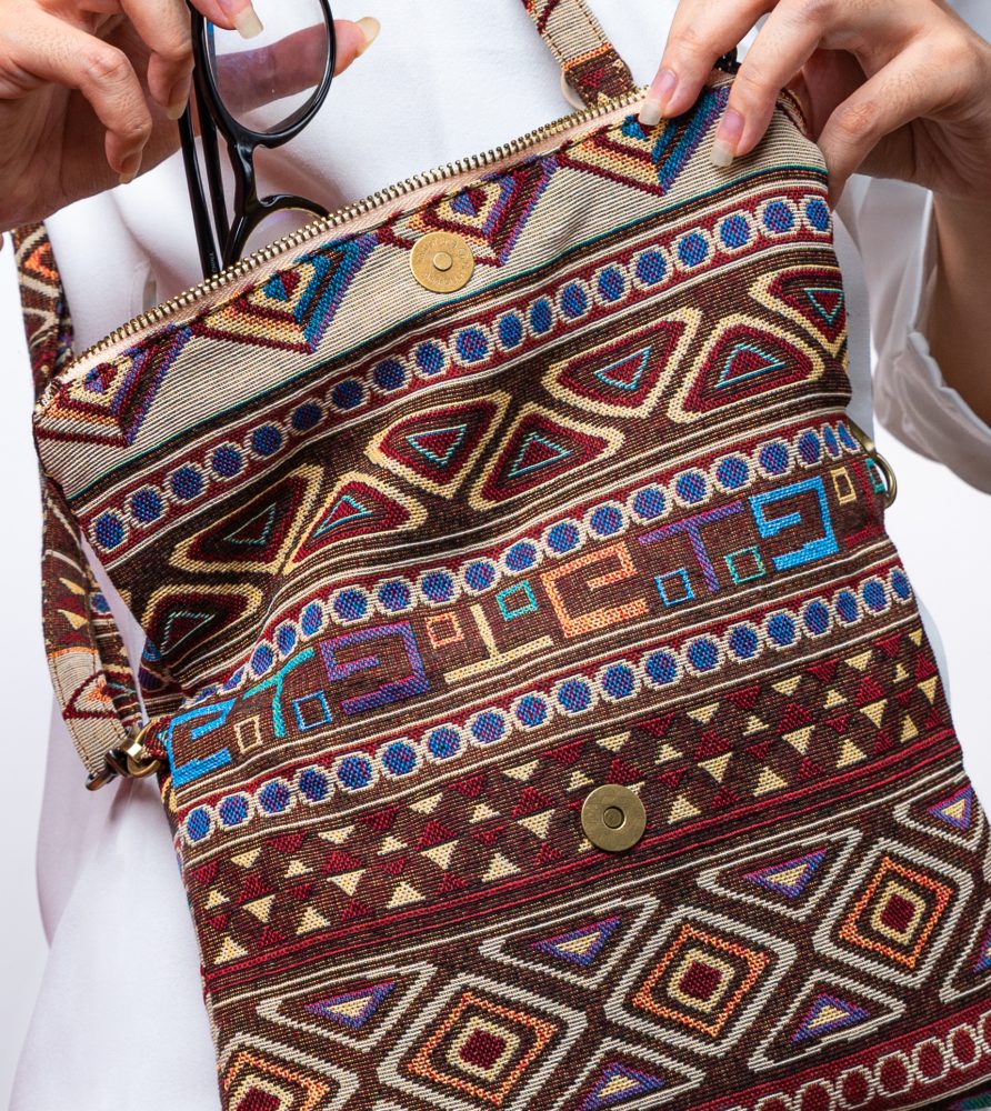 A stylish fold-over crossbody bag or shoulder bag made from a dark brown woven fabric. The bag features an intricate Southwestern or Aztec geometric pattern with horizontal stripes of diamond, zigzag, and Greek key motifs in shades of burgundy, deep blue, turquoise, and orange. The bag has a small zipper along the fold and a wide, matching patterned fabric strap with silver-toned hardware, showcasing an eclectic fashion accessory for everyday wear or travel.