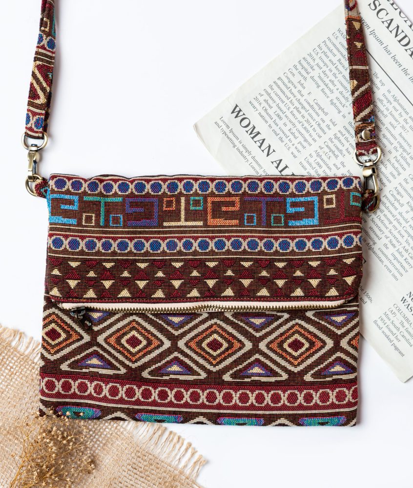 A stylish fold-over crossbody bag or shoulder bag made from a dark brown woven fabric. The bag features an intricate Southwestern or Aztec geometric pattern with horizontal stripes of diamond, zigzag, and Greek key motifs in shades of burgundy, deep blue, turquoise, and orange. The bag has a small zipper along the fold and a wide, matching patterned fabric strap with silver-toned hardware, showcasing an eclectic fashion accessory for everyday wear or travel.