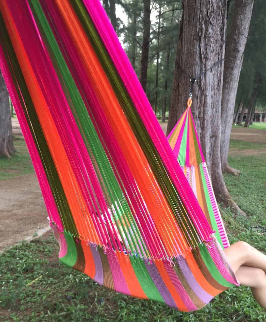 a girl resting in a bright pink and orange striped hammock set up between two trees in a grassy park.