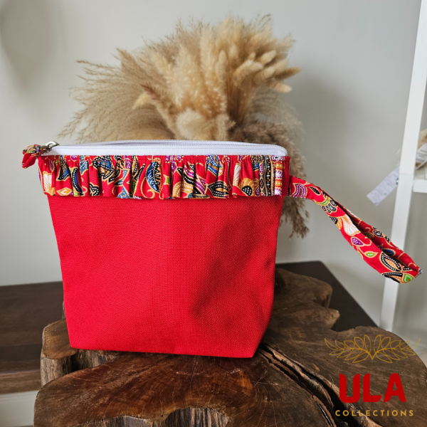 A vibrant zippered pouch bag or clutch with a solid red cotton body. The bag is decorated with a wide, ruffled trim and interior lining made from a red batik fabric with a multicolor floral print in shades of gold, teal, and brown. The bag is shown open and includes a matching batik fabric wrist strap or pull tab. It is resting on a dark, rustic wooden surface, with a logo and dried pampas grass in the background, highlighting an eclectic fashion accessory perfect for travel or a statement handbag.