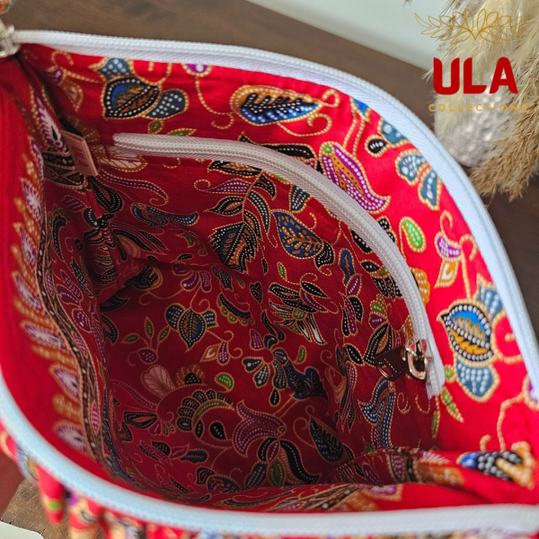 A vibrant zippered pouch bag or clutch with a solid red cotton body. The bag is decorated with a wide, ruffled trim and interior lining made from a red batik fabric with a multicolor floral print in shades of gold, teal, and brown. The bag is shown open and includes a matching batik fabric wrist strap or pull tab. It is resting on a dark, rustic wooden surface, with a logo and dried pampas grass in the background, highlighting an eclectic fashion accessory perfect for travel or a statement handbag.