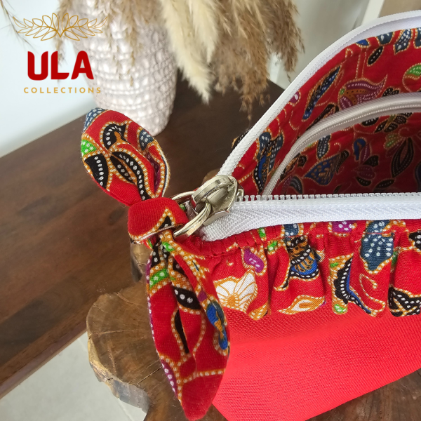 A vibrant zippered pouch bag or clutch with a solid red cotton body. The bag is decorated with a wide, ruffled trim and interior lining made from a red batik fabric with a multicolor floral print in shades of gold, teal, and brown. The bag is shown open and includes a matching batik fabric wrist strap or pull tab. It is resting on a dark, rustic wooden surface, with a logo and dried pampas grass in the background, highlighting an eclectic fashion accessory perfect for travel or a statement handbag.