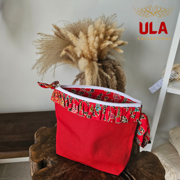 A vibrant zippered pouch bag or clutch with a solid red cotton body. The bag is decorated with a wide, ruffled trim and interior lining made from a red batik fabric with a multicolor floral print in shades of gold, teal, and brown. The bag is shown open and includes a matching batik fabric wrist strap or pull tab. It is resting on a dark, rustic wooden surface, with a logo and dried pampas grass in the background, highlighting an eclectic fashion accessory perfect for travel or a statement handbag.