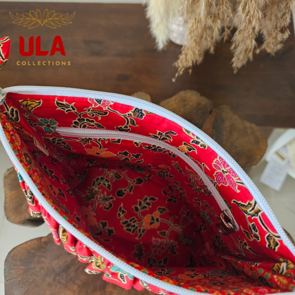 A vibrant zippered pouch bag or clutch with a solid red cotton body. The bag is decorated with a wide, ruffled trim and interior lining made from a red batik fabric with a multicolor floral print in shades of gold, teal, and brown. The bag is shown open and includes a matching batik fabric wrist strap or pull tab. It is resting on a dark, rustic wooden surface, with a logo and dried pampas grass in the background, highlighting an eclectic fashion accessory perfect for travel or a statement handbag.