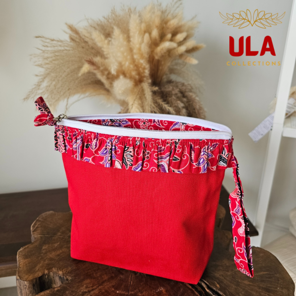 A vibrant zippered pouch bag or clutch with a solid bright red cotton body. The bag is decorated with a wide, ruffled trim and interior lining made from a red batik fabric with a multicolor floral print in shades of purple, white, and dark red. The bag is shown open and includes a matching batik fabric wrist strap. It rests on a dark, rustic wooden surface, with a logo and dried pampas grass in the background, highlighting an eclectic fashion accessory perfect for travel or a statement handbag.