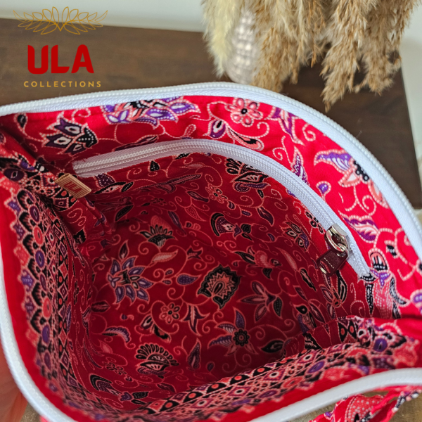 A vibrant zippered pouch bag or clutch with a solid bright red cotton body. The bag is decorated with a wide, ruffled trim and interior lining made from a red batik fabric with a multicolor floral print in shades of purple, white, and dark red. The bag is shown open and includes a matching batik fabric wrist strap. It rests on a dark, rustic wooden surface, with a logo and dried pampas grass in the background, highlighting an eclectic fashion accessory perfect for travel or a statement handbag.