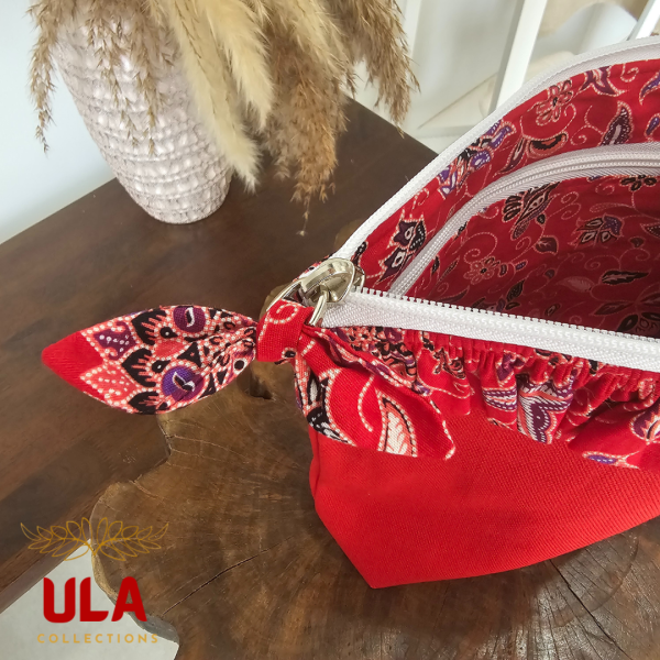 A vibrant zippered pouch bag or clutch with a solid bright red cotton body. The bag is decorated with a wide, ruffled trim and interior lining made from a red batik fabric with a multicolor floral print in shades of purple, white, and dark red. The bag is shown open and includes a matching batik fabric wrist strap. It rests on a dark, rustic wooden surface, with a logo and dried pampas grass in the background, highlighting an eclectic fashion accessory perfect for travel or a statement handbag.