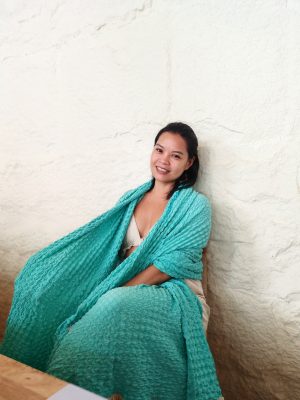 Portrait of a smiling woman wrapped in a vibrant turquoise blue textured shawl, sitting against a white stone wall.